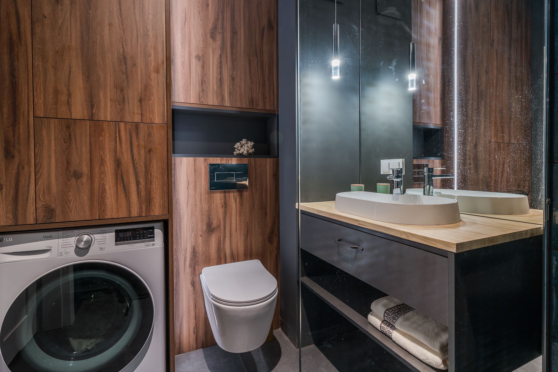 Nowoczesna łazienka z drewnianymi panelami, podwieszaną toaletą, pralką LG i umywalką nablatową na drewnianym blacie, oświetlona wiszącymi lampami.