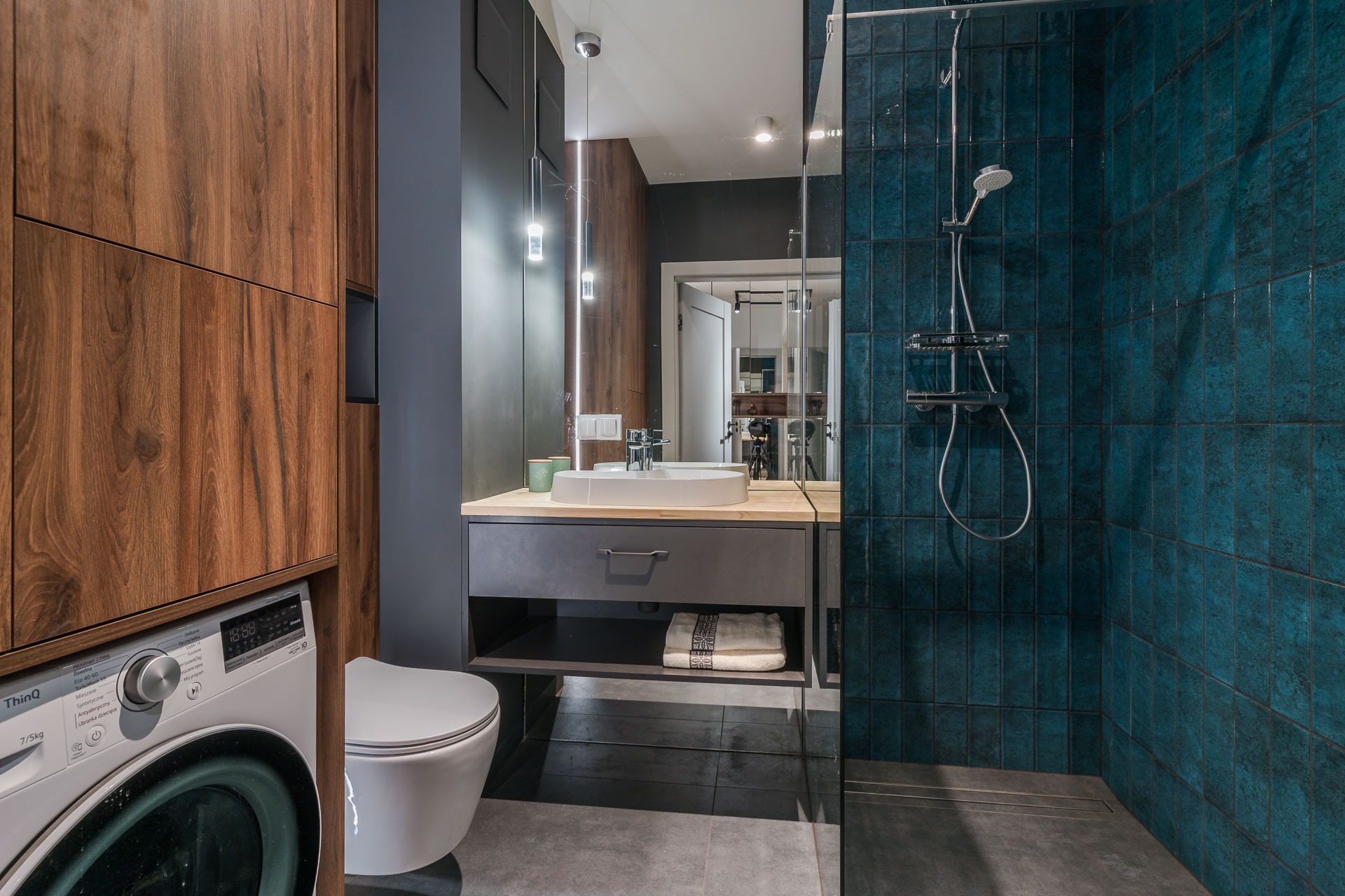 Nowoczesna łazienka z ciemnozielonymi płytkami w kabinie prysznicowej, umywalką na szafce z szufladą, toaletą i pralką ukrytą w drewnianej zabudowie.