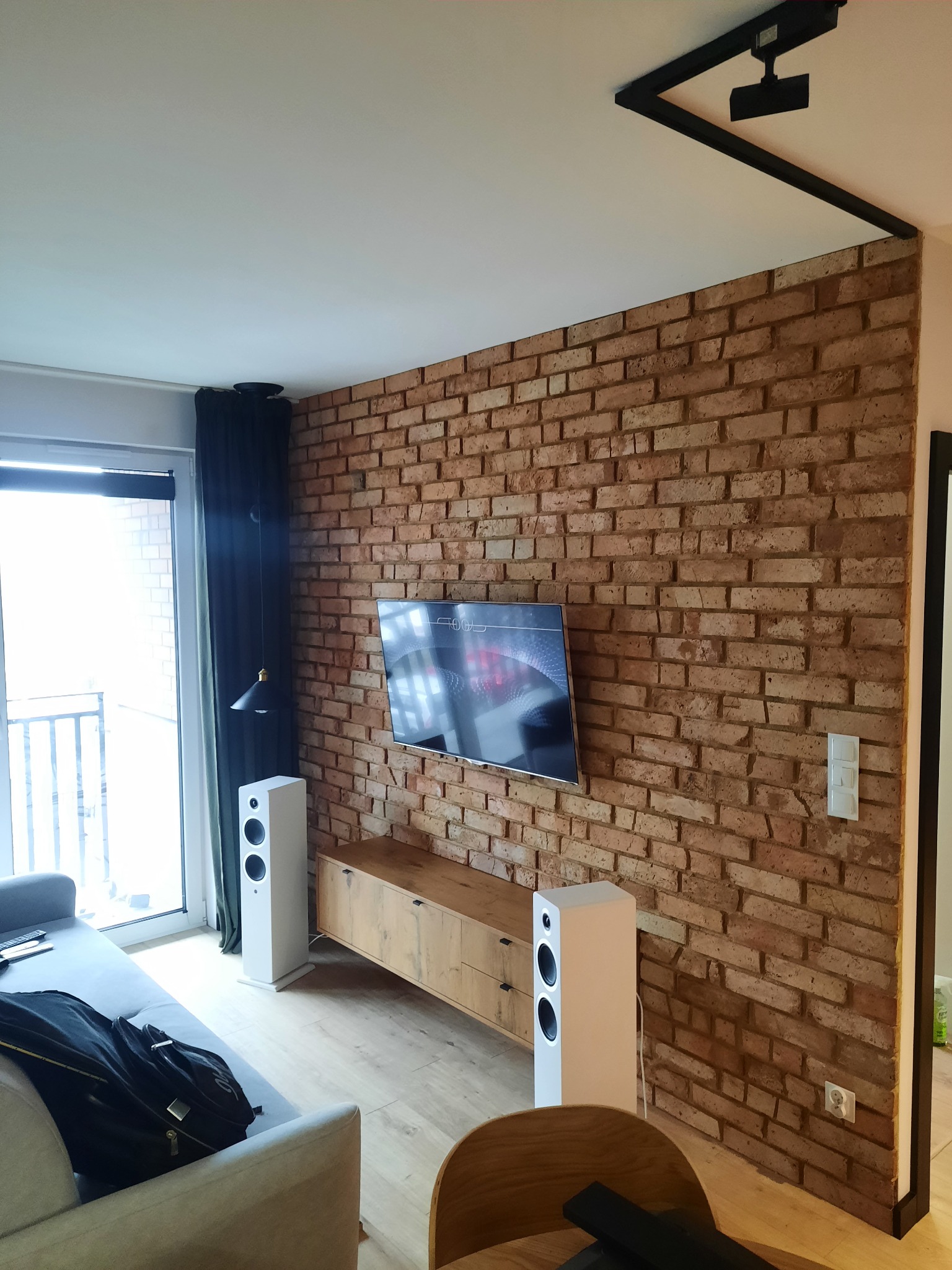 Salon z dekoracyjną ścianą z cegły, telewizorem, drewnianą szafką RTV i białymi głośnikami podświetlanymi przez lampę wiszącą przy balkonie.