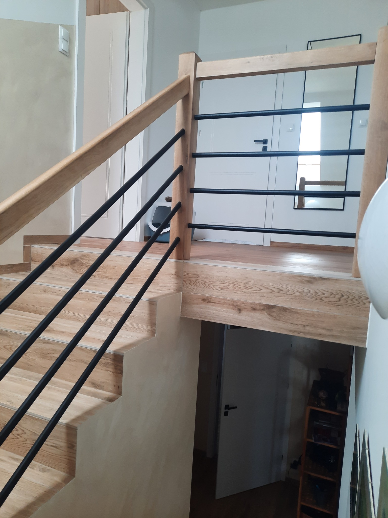 Nowoczesne schody drewniane z czarnymi, metalowymi balustradami, wykończone płytkami imitującymi drewno, widok z dołu na górny poziom z białymi drzwiami i lustrem.