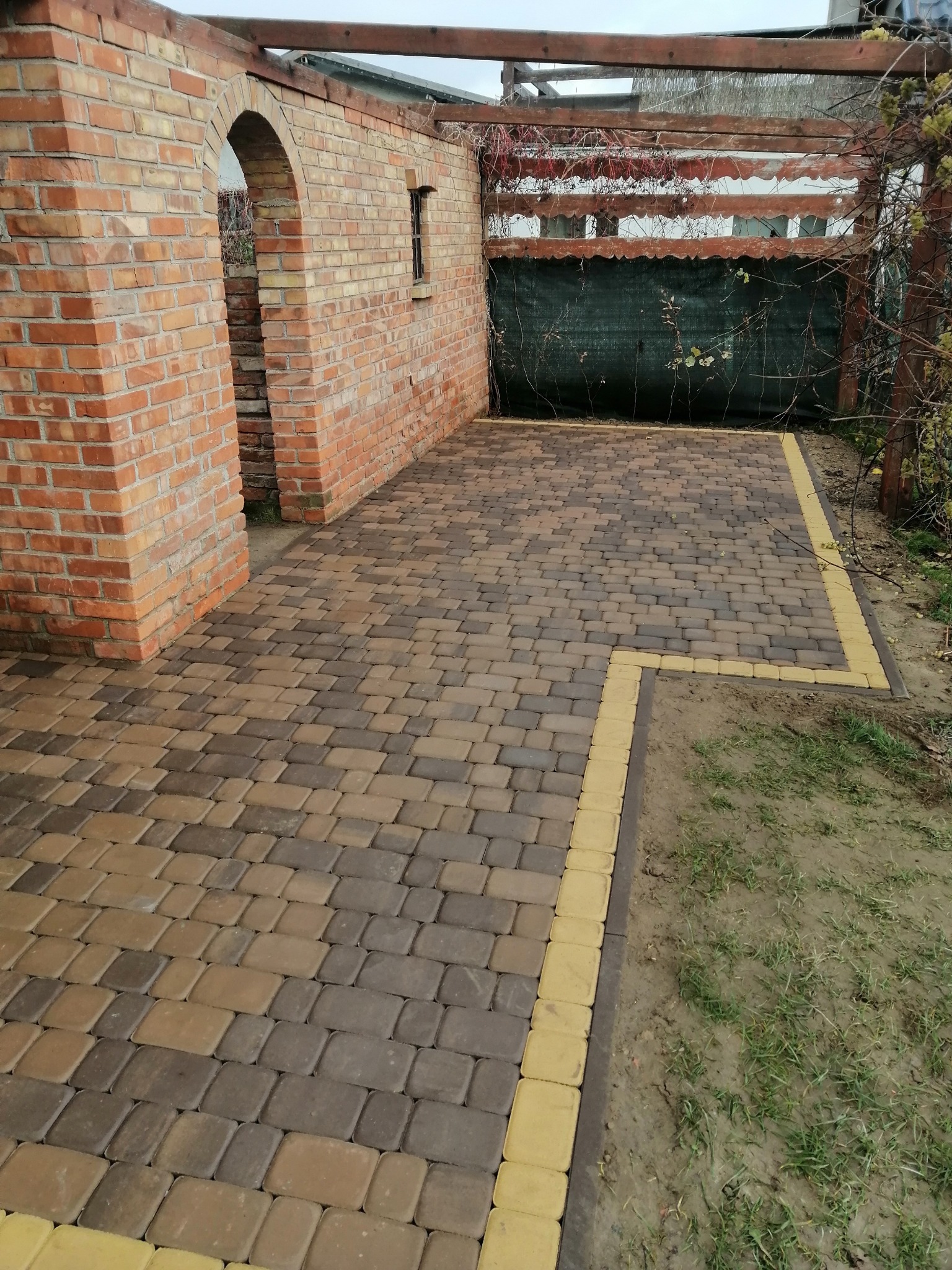 Ułożona kostka brukowa w odcieniach brązu i beżu z żółtymi obrzeżami na zewnątrz, z fragmentem ceglanego muru z łukowym przejściem i drewnianą konstrukcją z tyłu.