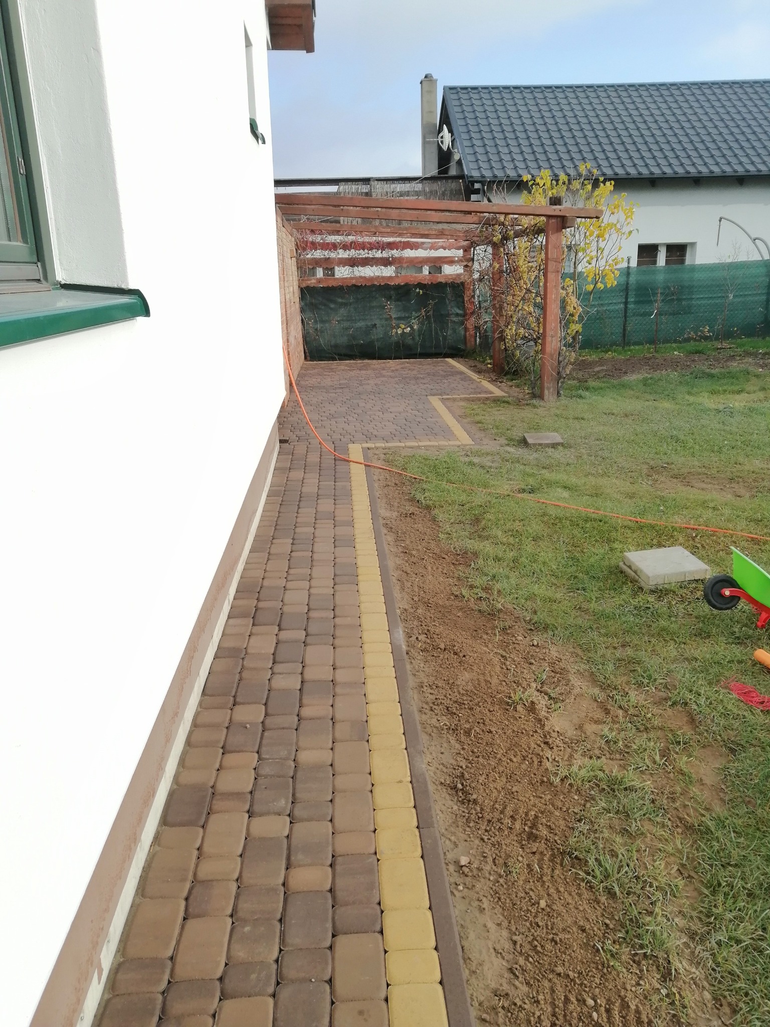 Ułożona kostka brukowa w dwóch odcieniach brązu i żółci przy ścianie budynku w Welk, widoczna pergola i trawnik.