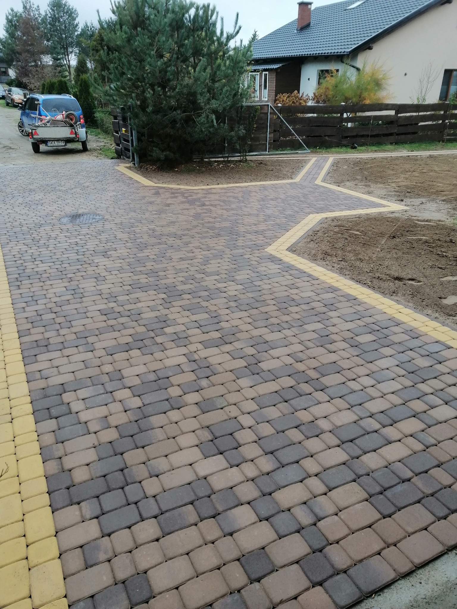 Ułożona kostka brukowa w odcieniach szarości i brązu z żółtymi obrzeżami, tworząca ścieżkę na posesji z widocznym samochodem z przyczepką w tle.