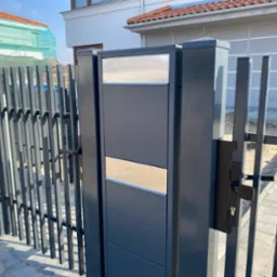 Nowoczesna, grafitowa brama wjazdowa z metalowymi prętami, skrzynką na listy z elementami ze stali nierdzewnej i czarną klamką, na tle jasnej elewacji domu.