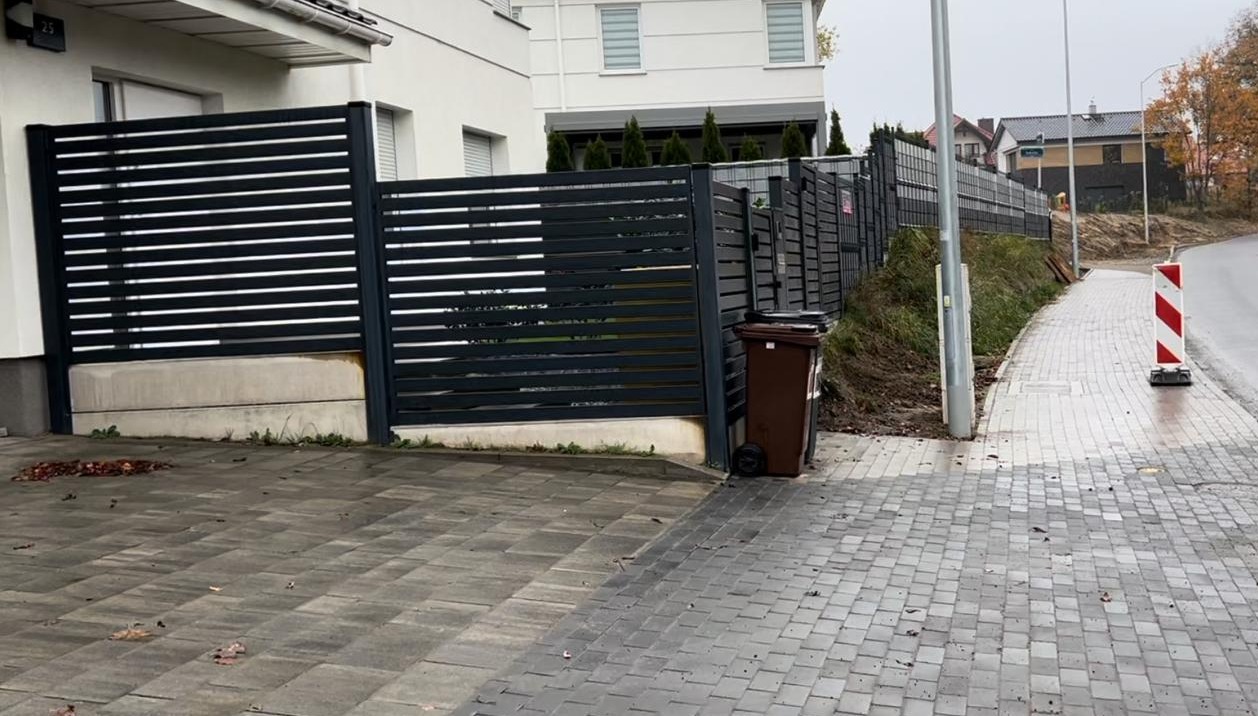 Nowoczesne, poziome ogrodzenie z czarnych paneli zamontowane na betonowej podmurówce, widoczne z zewnątrz posesji w pochmurny dzień. Obok stoi brązowy pojemnik na śmieci na kółkach, a w tle widać...