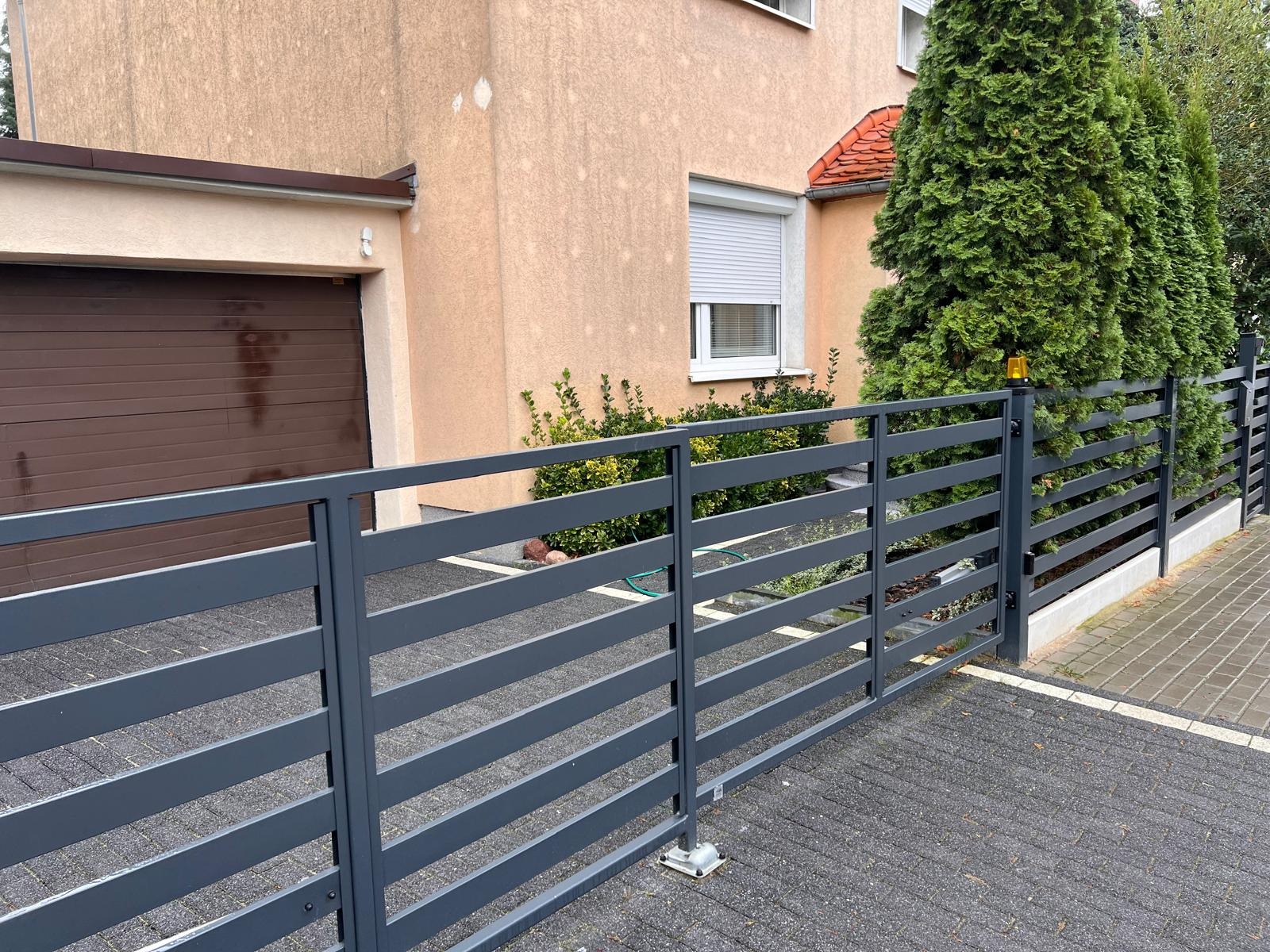 Nowoczesne, poziome ogrodzenie panelowe w kolorze grafitowym z bramą wjazdową, zamontowane na posesji z kostki brukowej, widoczne tuje i elewacja domu w tle.