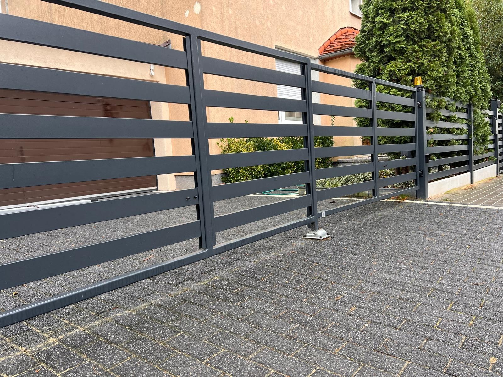 Nowoczesna, pozioma przesuwna brama z metalowych paneli w kolorze grafitowym, z widocznym mechanizmem automatycznego otwierania, na tle kostki brukowej i fragmentu domu z brązową bramą garażową.