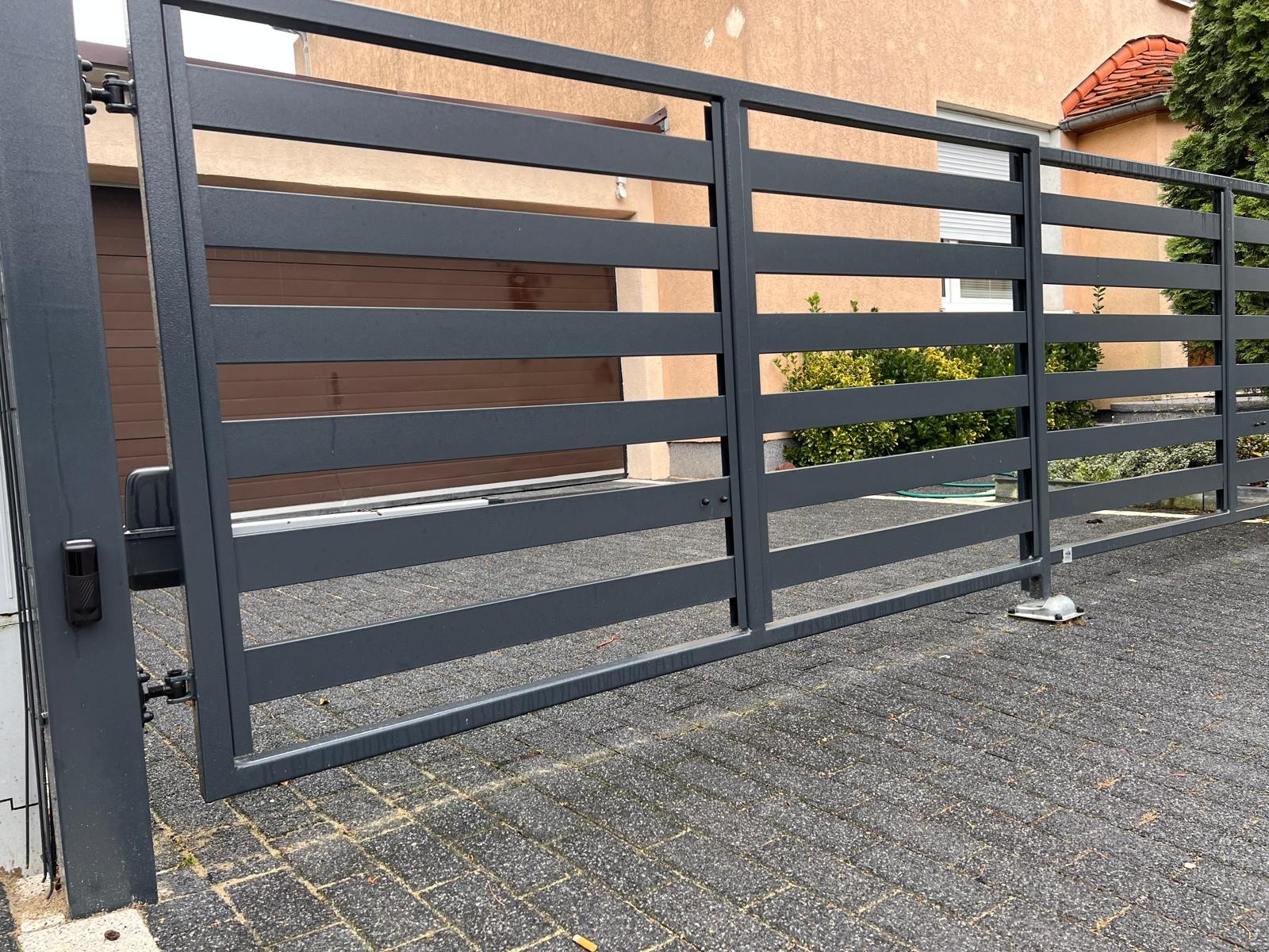 Nowoczesna brama wjazdowa przesuwna z szarych, poziomych profili stalowych, widoczny automat do bramy i czujnik ruchu, na pierwszym planie kostka brukowa.