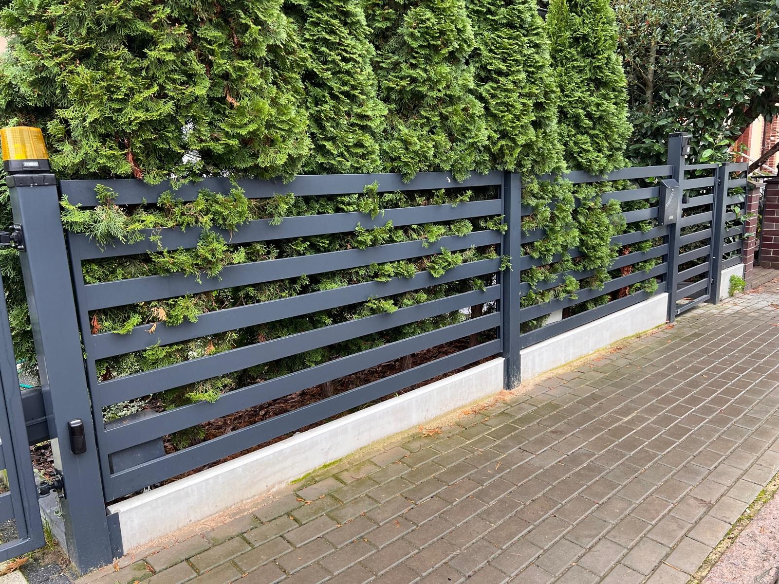 Nowoczesne, poziome przęsła w grafitowym kolorze, częściowo przesłonięte zielonymi tujami, tworzące minimalistyczne ogrodzenie posesji z kostki brukowej.