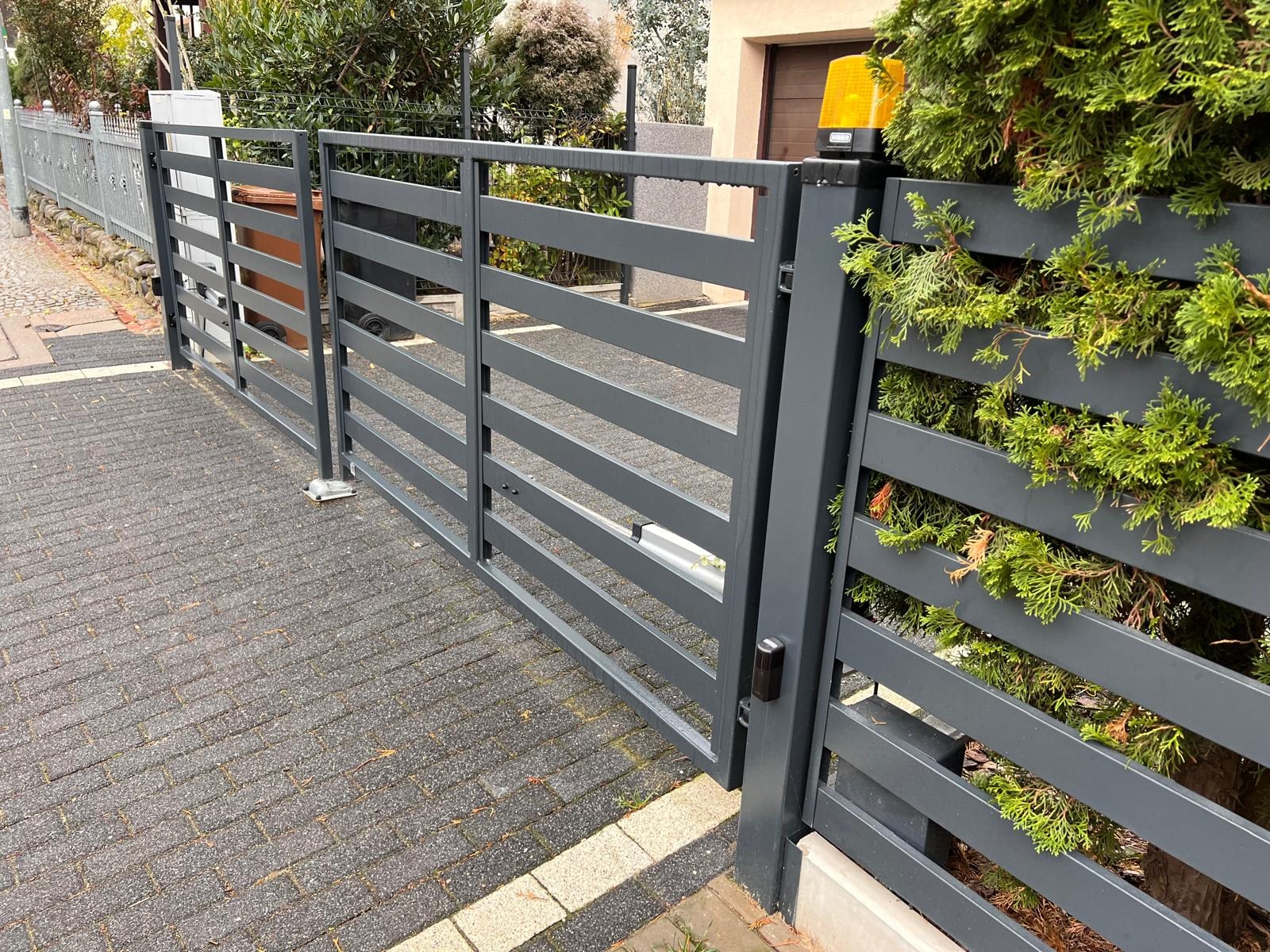 Nowoczesna, pozioma brama wjazdowa w kolorze grafitowym, z automatyką i lampą ostrzegawczą, zamontowana na posesji z brukowaną nawierzchnią i zielenią.