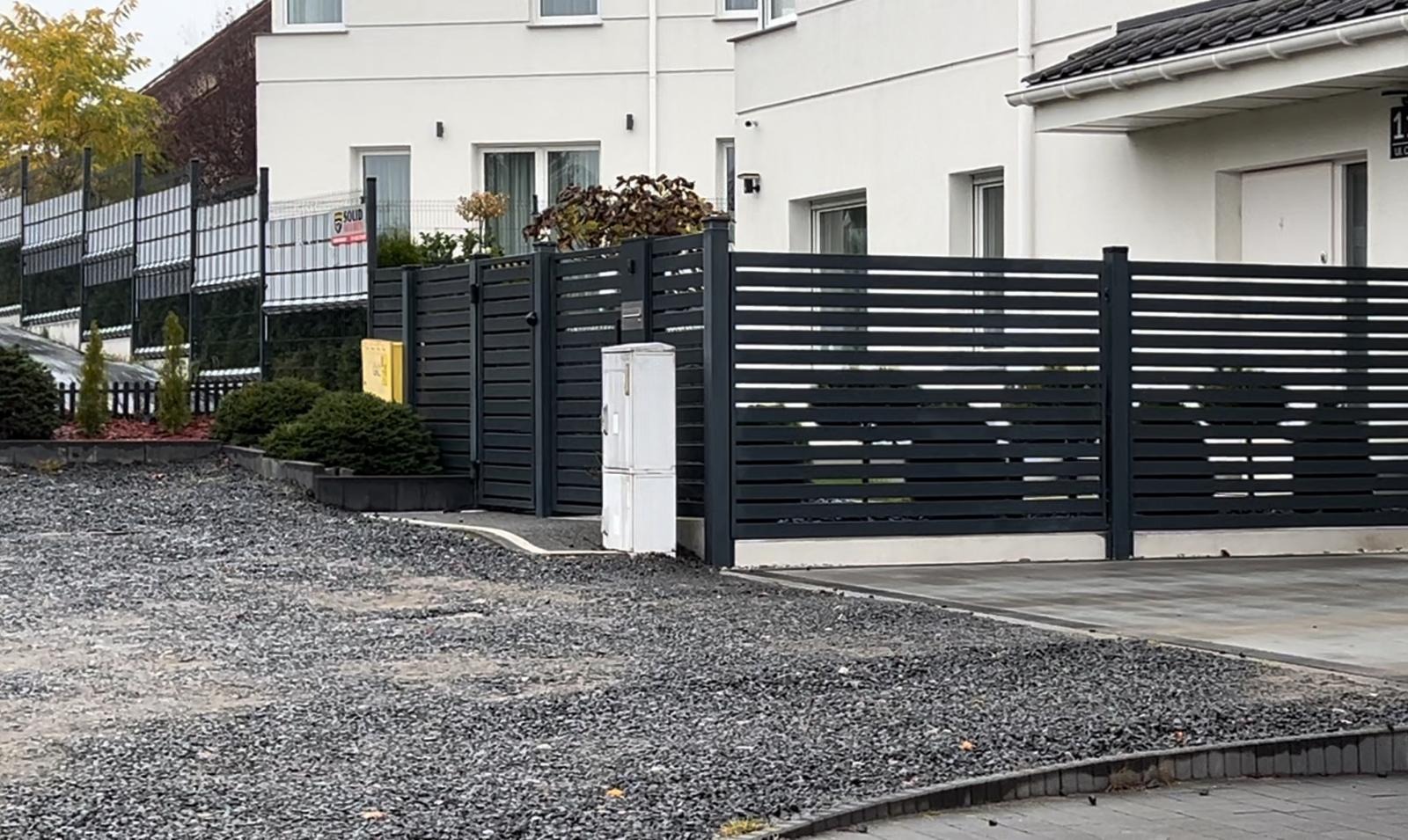 Nowoczesne ogrodzenie panelowe z poziomych, grafitowych listew, z widoczną skrzynką elektryczną i częścią furtki, na tle białego domu z szarym dachem i żwirowego podłoża.