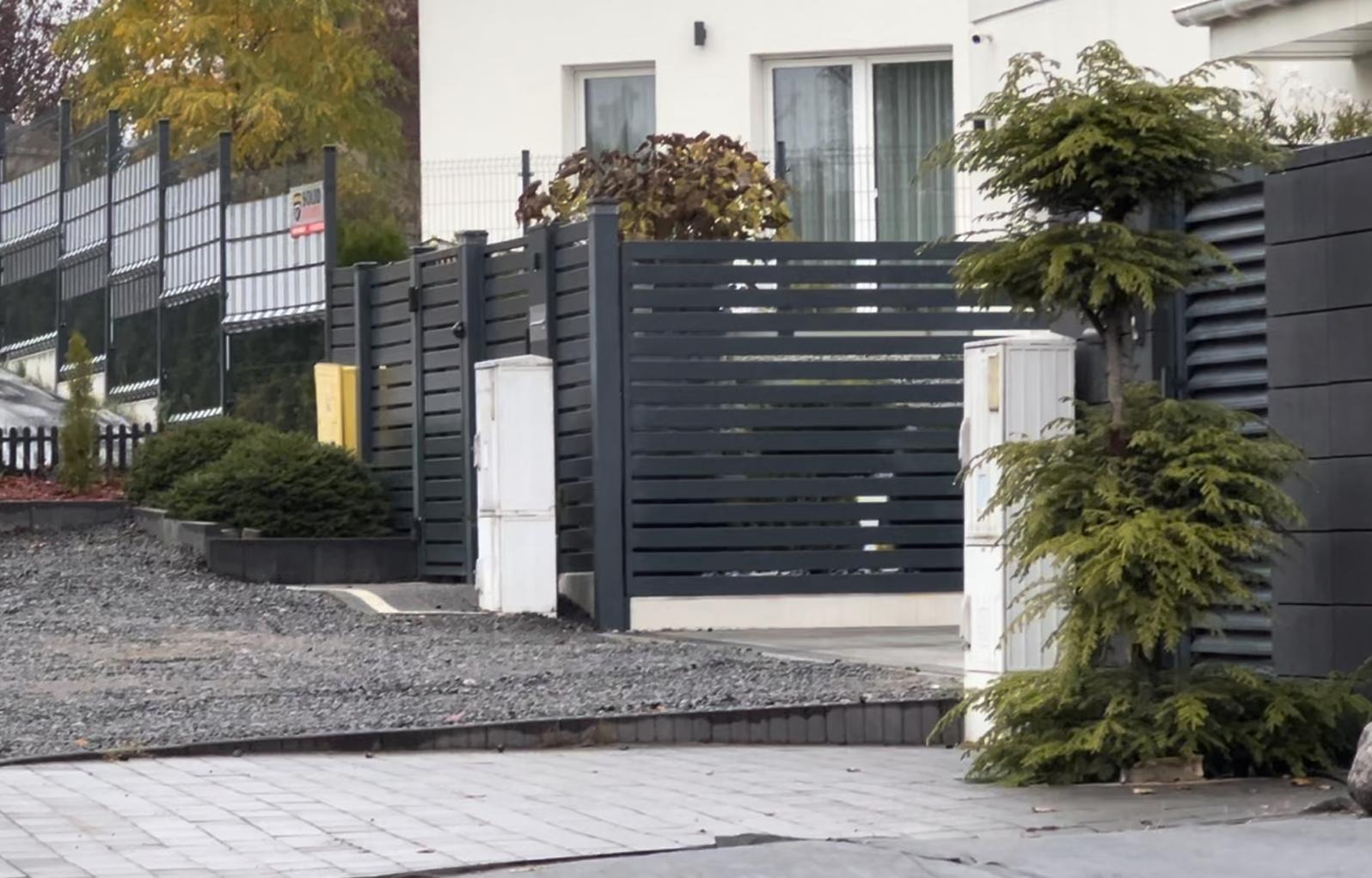 Nowoczesne ogrodzenie posesji z poziomych, grafitowych paneli, brama wjazdowa z tego samego materiału, na pierwszym planie ozdobny krzew iglasty formowany w kulę.