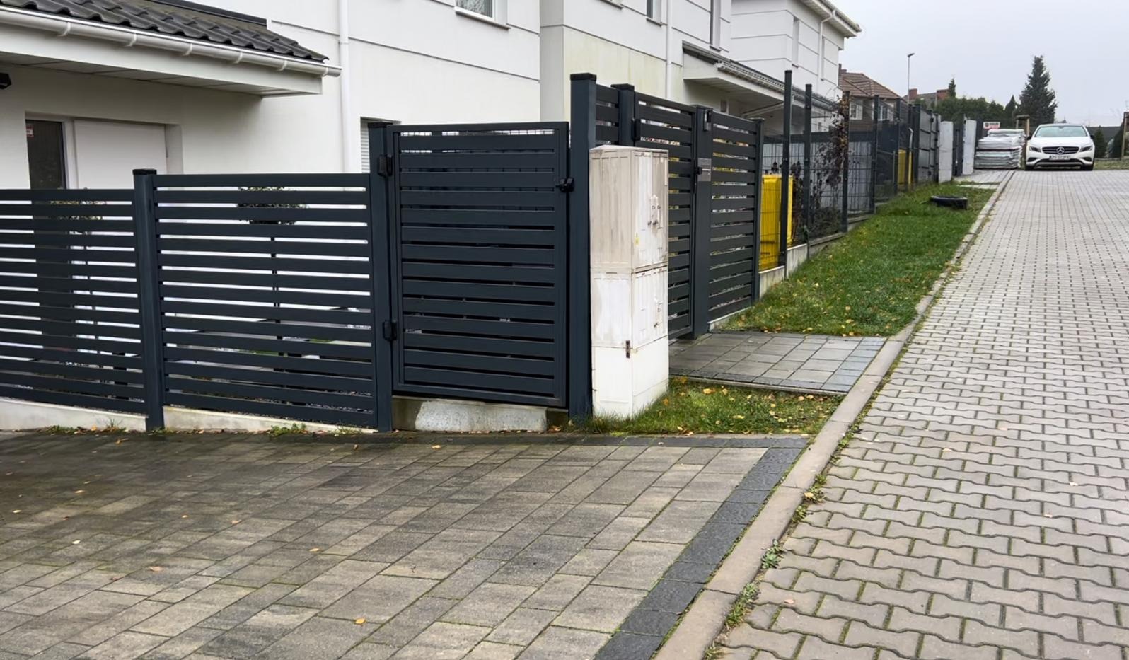 Nowoczesne, poziome, szare ogrodzenie z metalowych paneli z bramką, widok z zewnątrz na kostkę brukową i fragment domu.