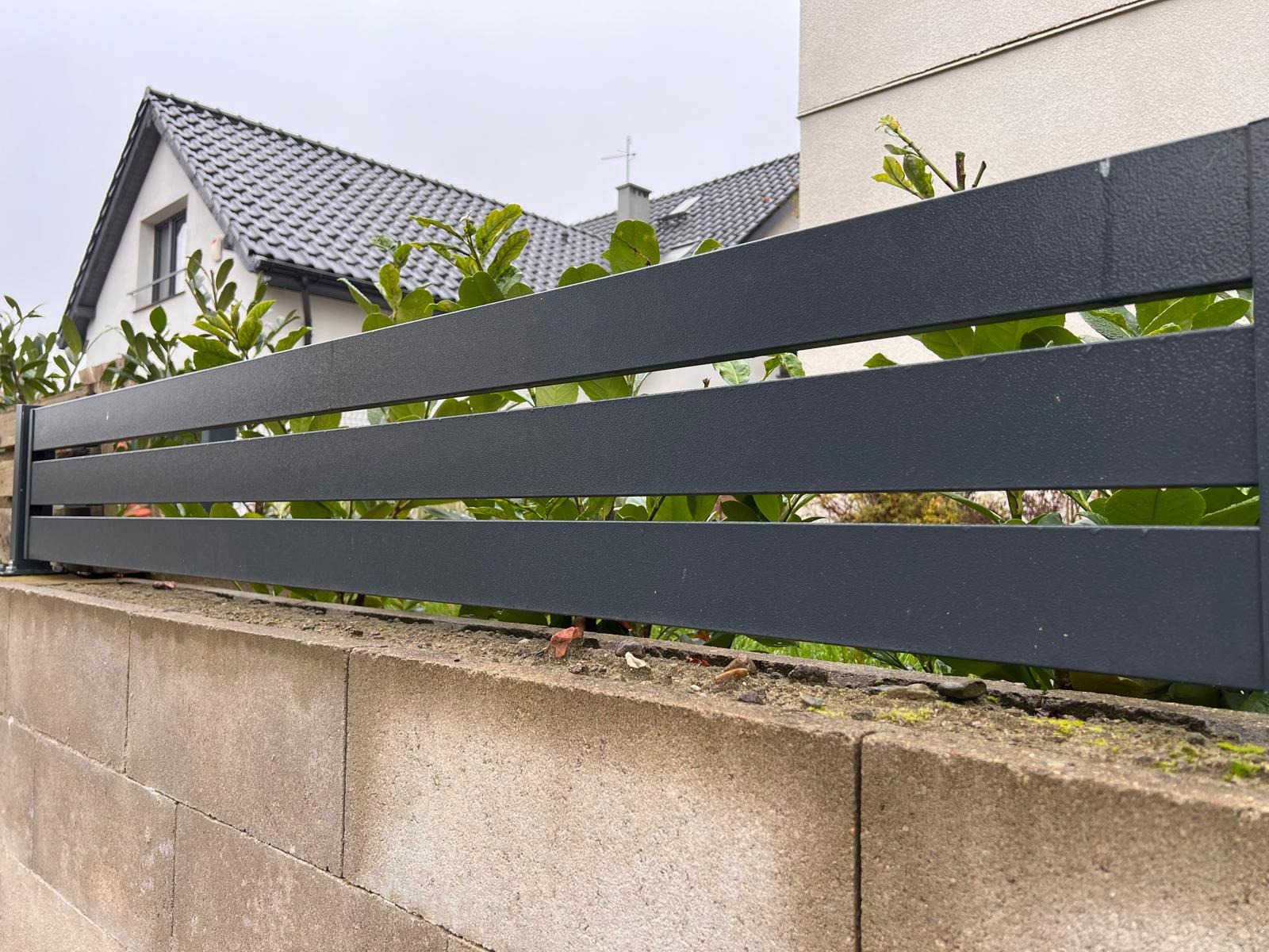 Grafitowe, poziome przęsła ogrodzenia na betonowej podmurówce, z zielenią w tle i fragmentem domu z szarą dachówką.