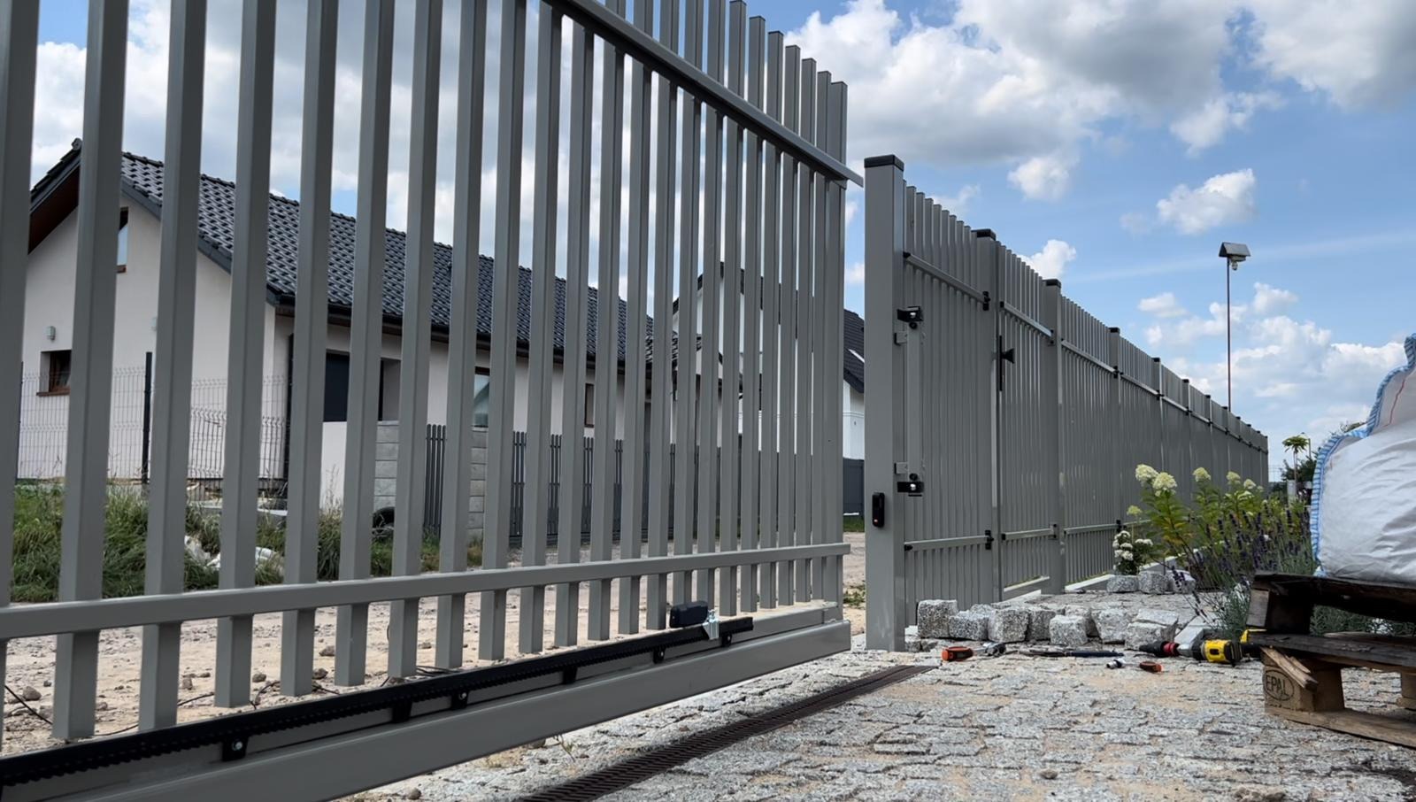 Otwarta, automatyczna brama wjazdowa z szarego metalu z pionowymi profilami, widoczny mechanizm napędowy, kostka brukowa na podjeździe, w tle dom jednorodzinny i błękitne niebo z chmurami.