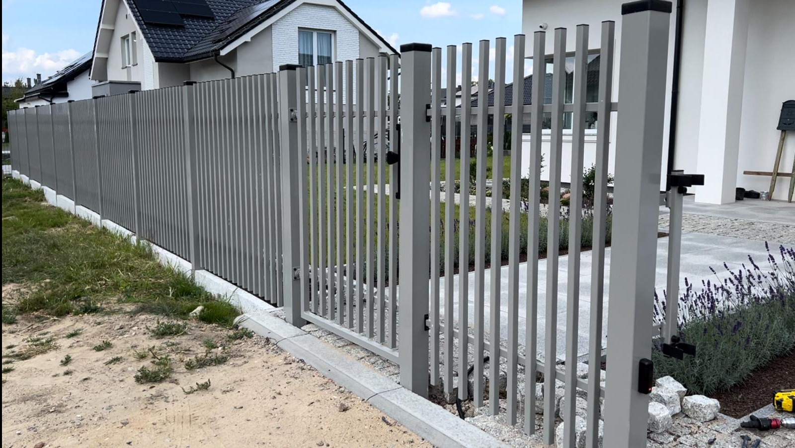 Nowoczesne ogrodzenie panelowe w kolorze szarym z bramą wjazdową, widoczne z perspektywy gruntu, z domem w tle i fragmentem rabaty z lawendą.
