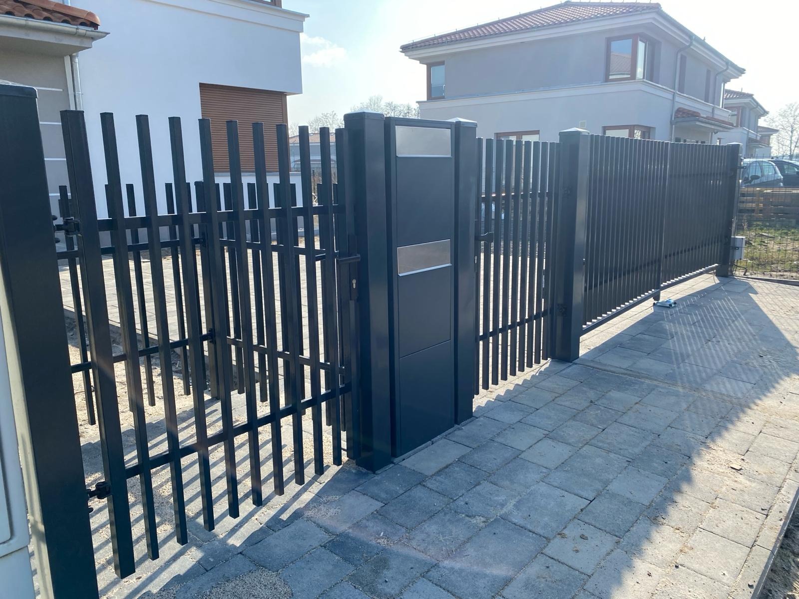 Nowoczesna, antracytowa brama wjazdowa z pionowych profili, z widocznymi elementami automatyki i skrzynką na listy, na tle kostki brukowej i zabudowy jednorodzinnej.