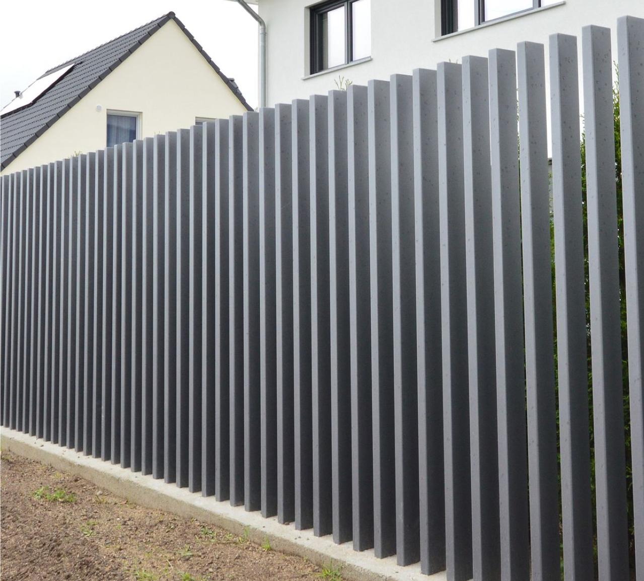 Nowoczesne ogrodzenie z pionowych, szarych profili metalowych, osadzone na betonowej podmurówce, z widocznymi budynkami w tle.