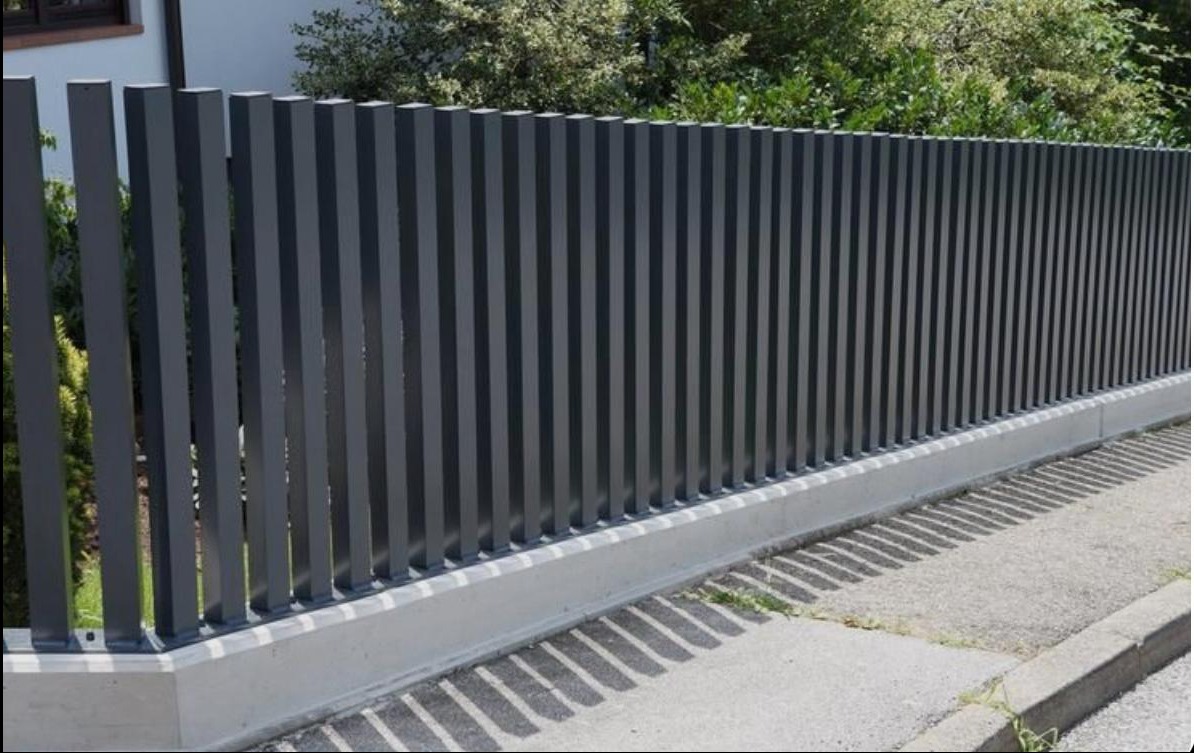 Nowoczesne, grafitowe ogrodzenie z pionowych, kwadratowych profili, osadzone na betonowej podmurówce, rzucające cienie na chodnik.