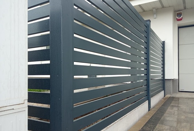 Nowoczesne, poziome przęsła ogrodzeniowe z metalu w kolorze grafitowym, widoczne krople deszczu, na tle białej ściany budynku z szarą podmurówką.