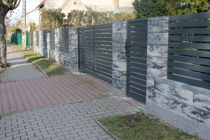 Nowoczesne ogrodzenie z przesuwną bramą w kolorze grafitowym, z elementami murowanymi z szarego kamienia o wyraźnym wzorze, widoczne z poziomu chodnika z kostki brukowej w słoneczny dzień.