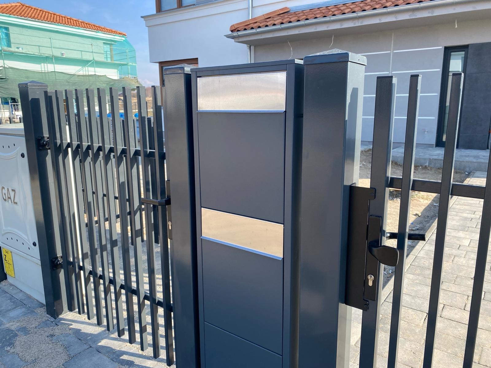 Nowoczesna, grafitowa brama wjazdowa z metalowymi prętami, skrzynką na listy z elementami ze stali nierdzewnej i czarną klamką, na tle jasnej elewacji domu.