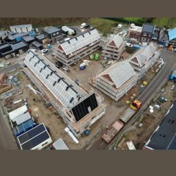 Dawid Romaniuk - Widok z drona na plac budowy z kilkoma domami w trakcie krycia dachu panelami słonecznymi, otoczonymi rusztowaniami i sprzętem budowlanym.