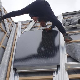 Dawid Romaniuk - Akrobata na dachu: mężczyzna w pozycji szpagatu w trakcie montażu panelu słonecznego, trzyma wiertarkę, widok z dołu na dach z drewnianą konstrukcją i częściowo ułożoną papą.