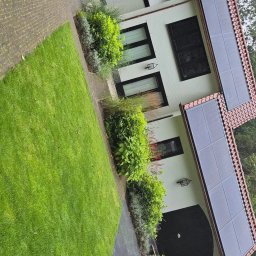 Dawid Romaniuk - Biały dom z czerwonym dachem i panelami słonecznymi, otoczony zielonym trawnikiem i roślinnością, widziany z perspektywy ogrodu.