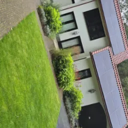 Biały dom z czerwonym dachem i panelami słonecznymi, otoczony zielonym trawnikiem i roślinnością, widziany z perspektywy ogrodu.