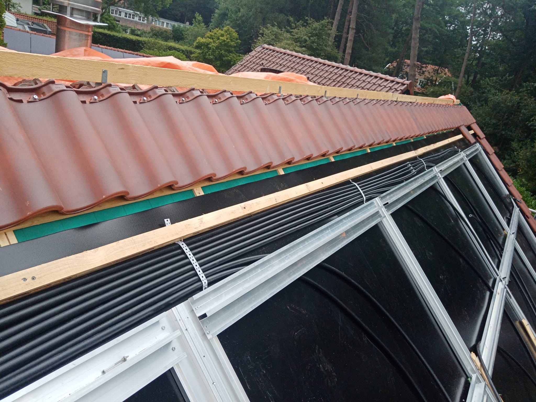 Montaż brązowej dachówki na drewnianej konstrukcji z widoczną membraną dachową, czarną izolacją i metalowym stelażem, w tle drzewa i budynki.