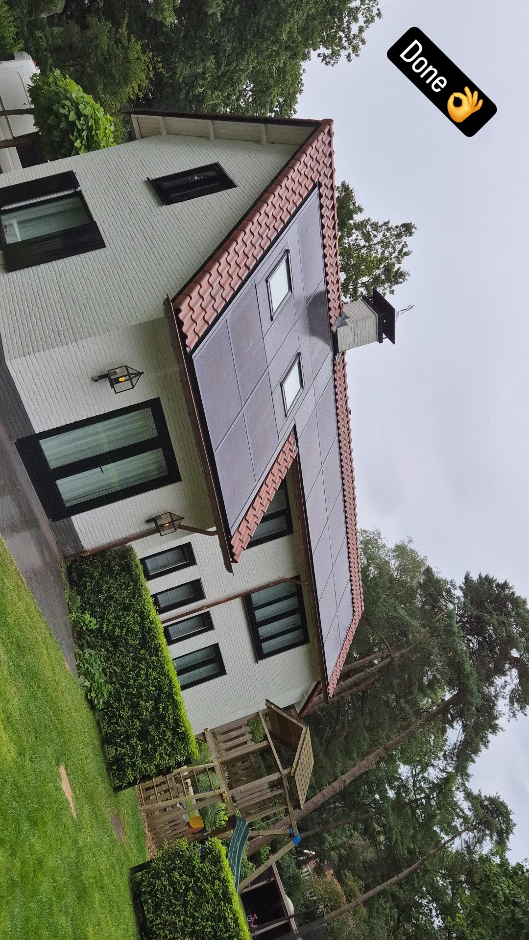 Biały dom jednorodzinny z nową, ciemną dachówką ceramiczną i oknami dachowymi, otoczony zielenią, widoczny z poziomu trawnika w pochmurny dzień. Na dachu widoczna naklejka 'Done'.