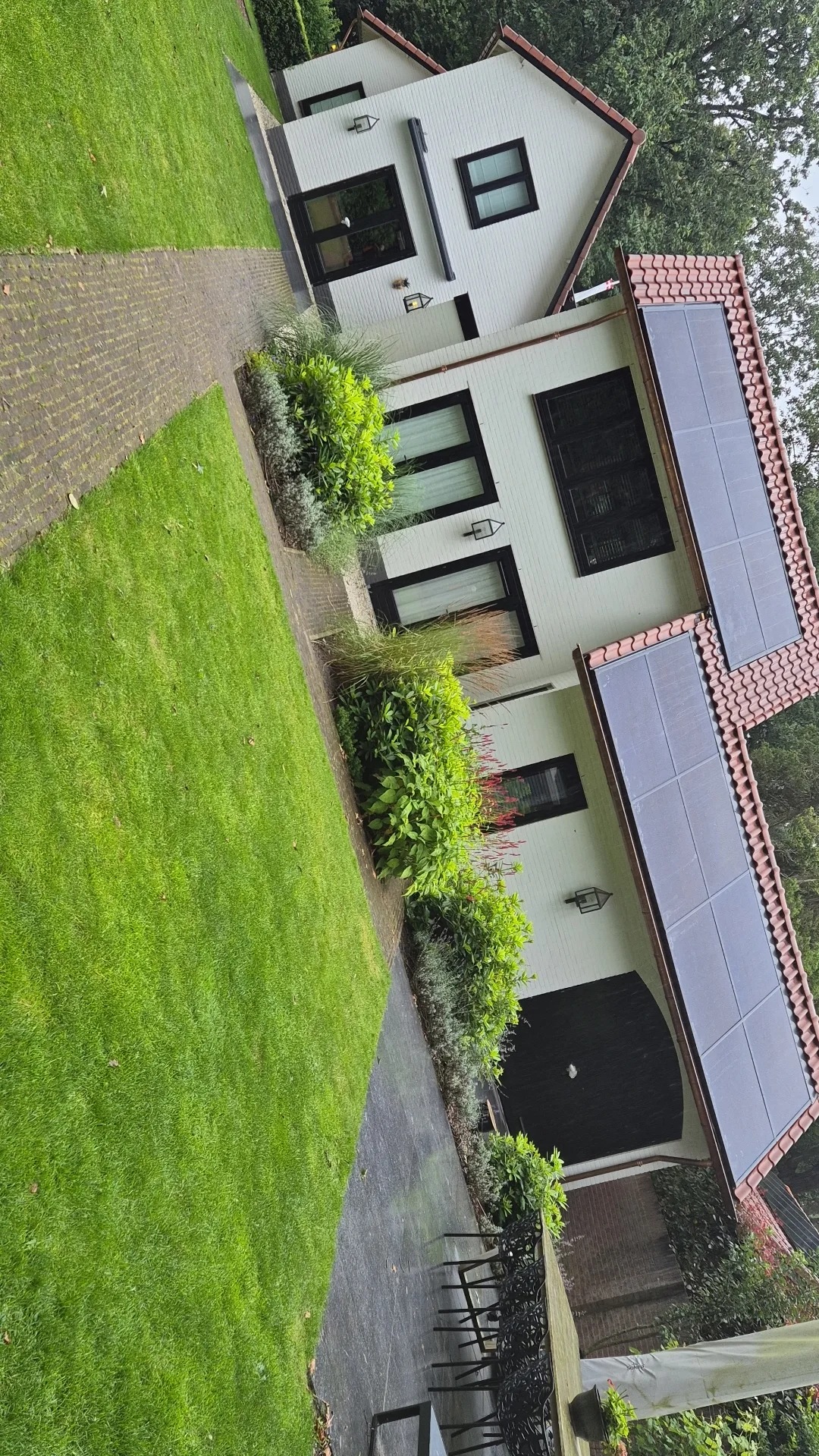 Biały dom z czerwonym dachem i panelami słonecznymi, otoczony zielonym trawnikiem i roślinnością, widziany z perspektywy ogrodu.