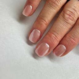 COSMETICA ROSA TERESA KUCHCIŃSKA - Dłoń z delikatnym, błyszczącym manicure hybrydowym w odcieniach nude na krótkich, kwadratowych paznokciach, z pierścionkiem na palcu wskazującym, na białym, teksturowanym tle.