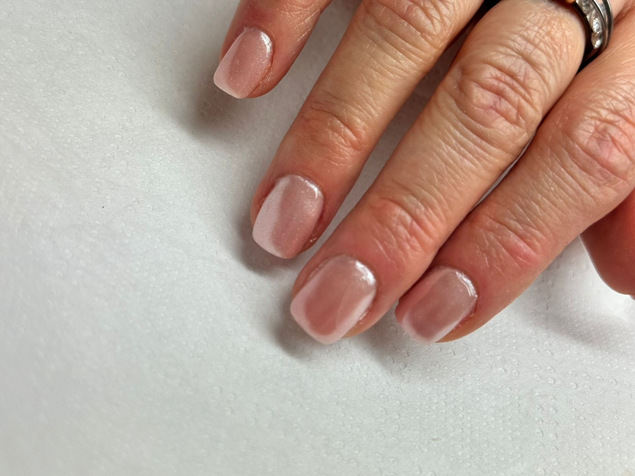 Dłoń z delikatnym, błyszczącym manicure hybrydowym w odcieniach nude na krótkich, kwadratowych paznokciach, z pierścionkiem na palcu wskazującym, na białym, teksturowanym tle.