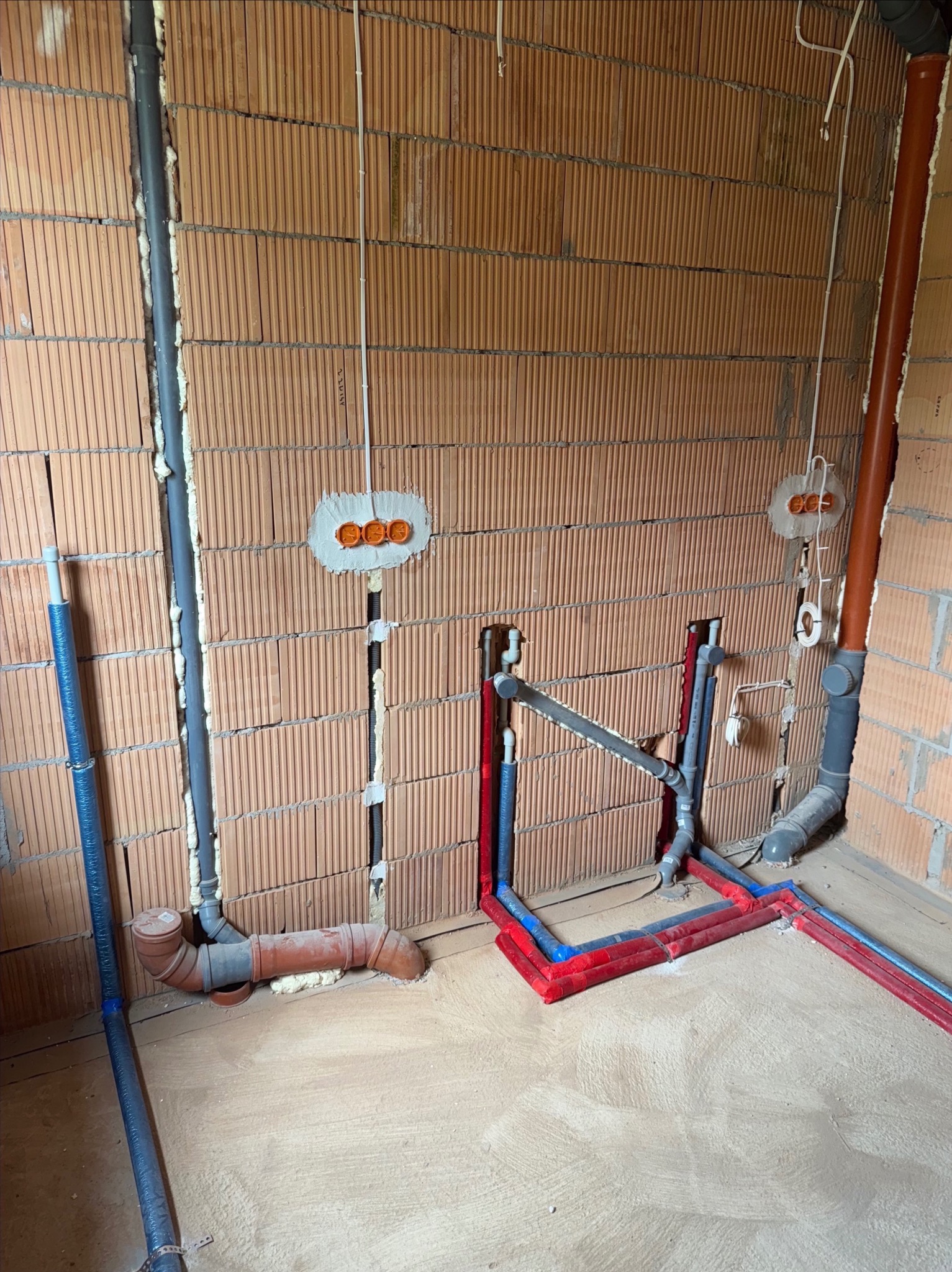 Instalacja wodno-kanalizacyjna w surowym wnętrzu: rury w kolorach czerwonym i niebieskim ułożone na podłodze, pionowe szare rury przy ścianach z cegły.