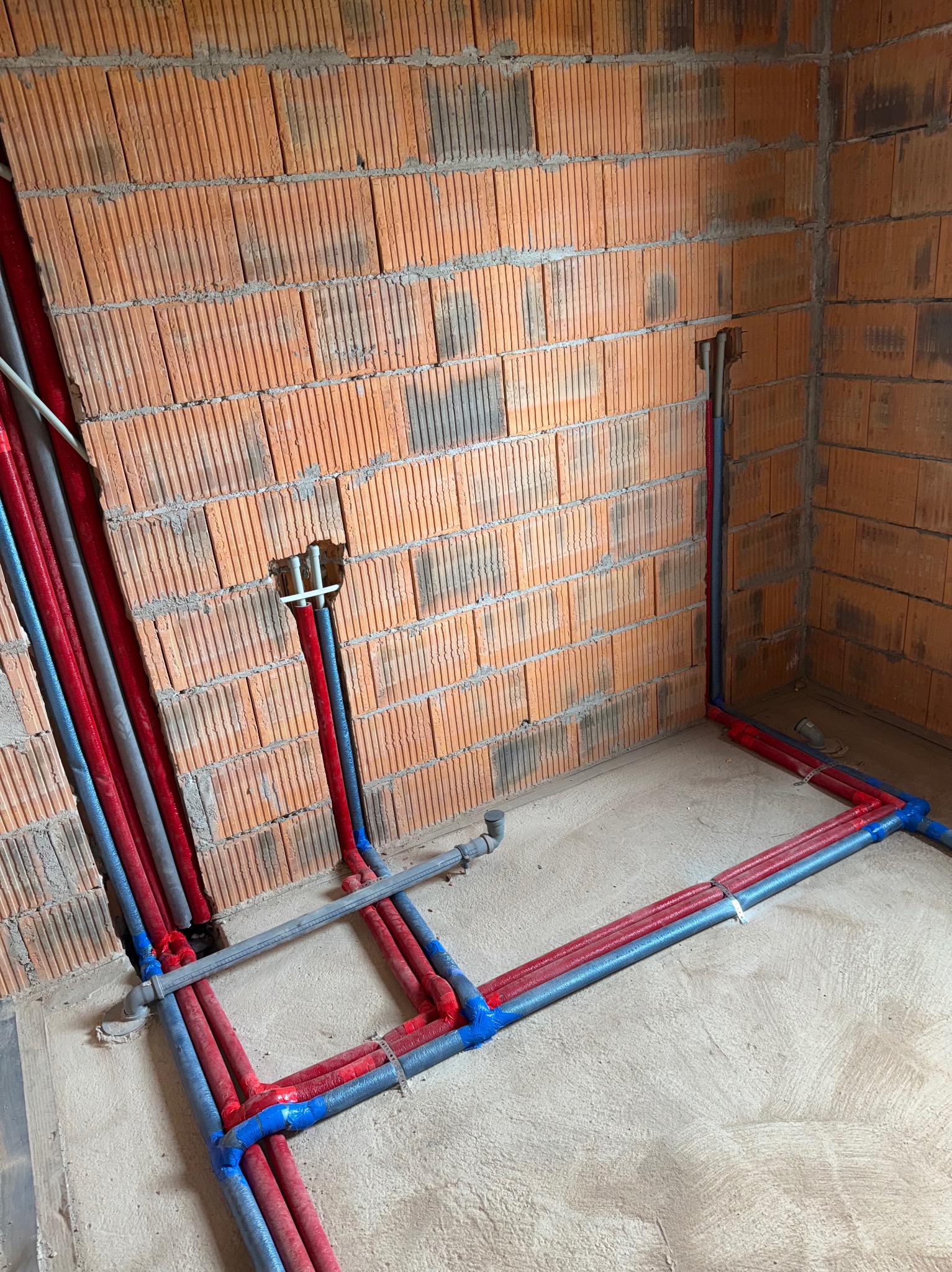 Instalacja wodna i grzewcza w surowym wnętrzu: czerwone i szare rury prowadzone wzdłuż ceglanej ściany i podłogi, widoczne mocowania i fragmenty instalacji w ścianie.