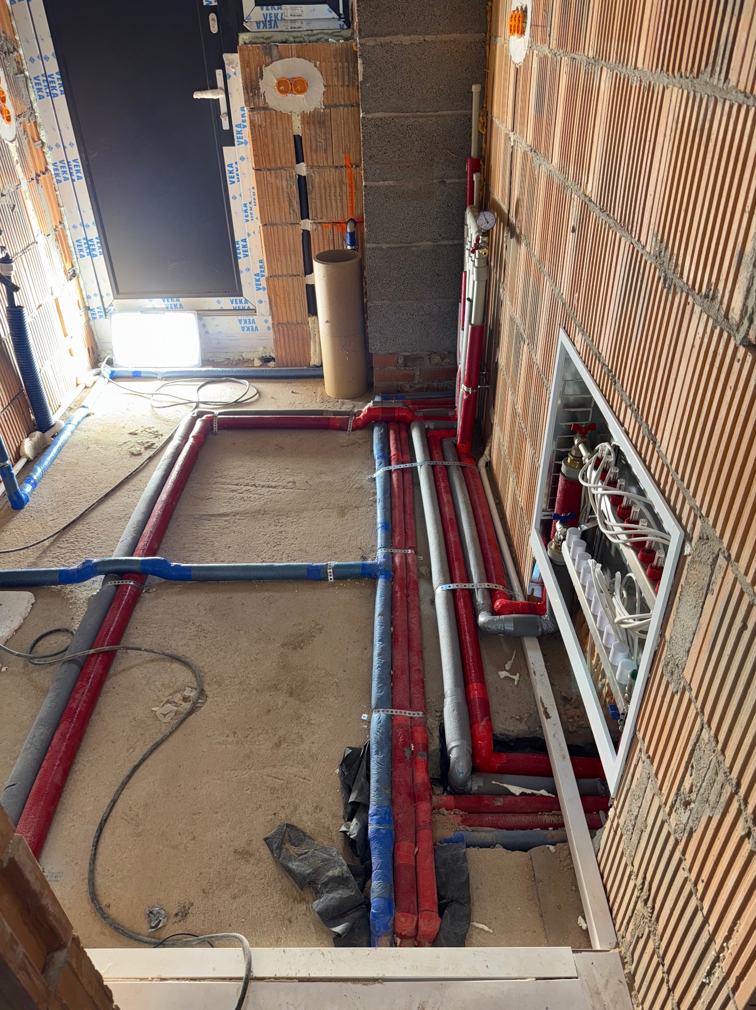 Instalacja ogrzewania podłogowego w surowym stanie: rury grzewcze w izolacji, rozdzielacz wnękowy, ściany z cegły dziurawki, okno z profili VEKA.