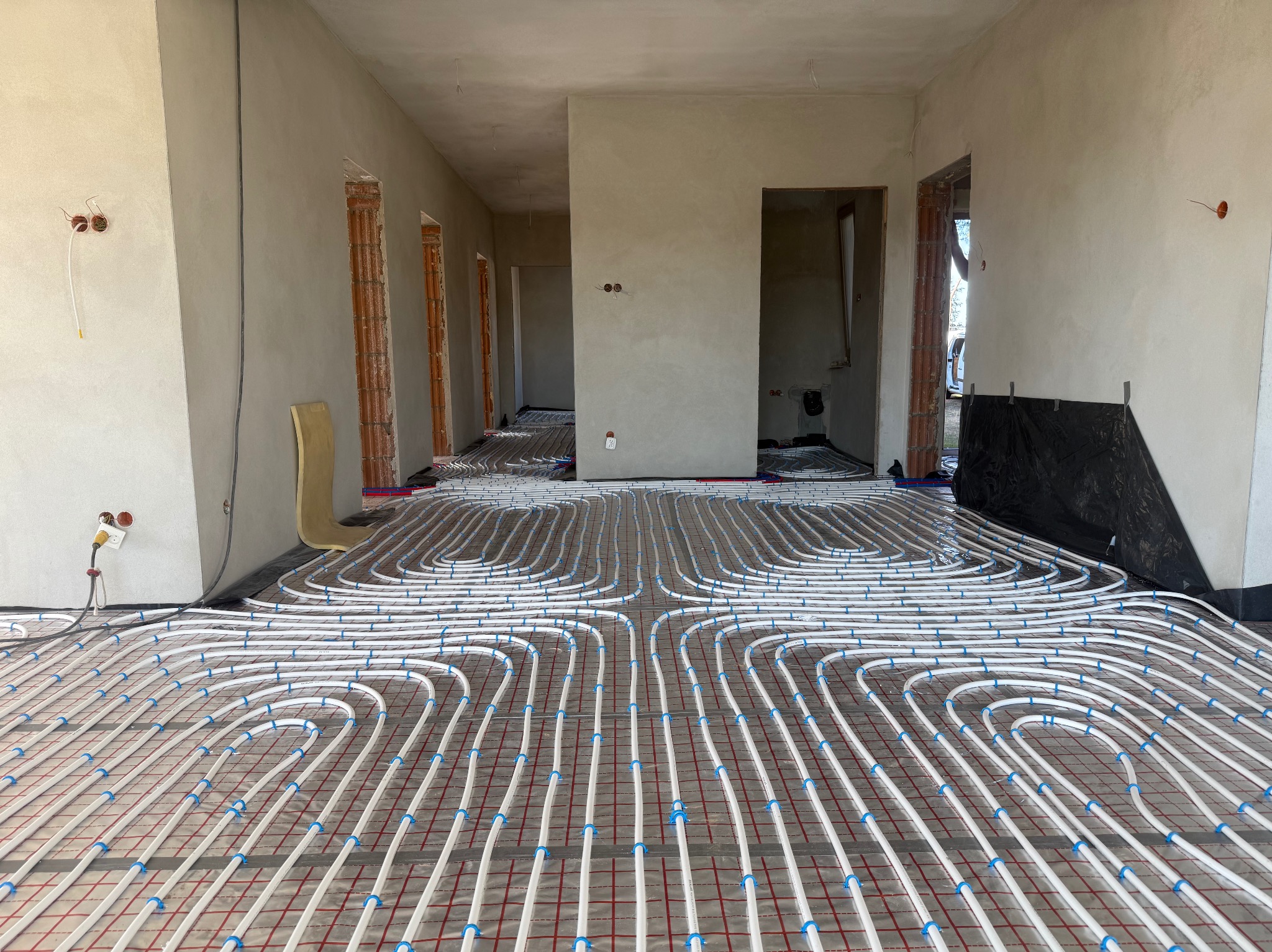 Białe rury ogrzewania podłogowego, mocowane niebieskimi uchwytami, ułożone na czerwonej siatce w surowym wnętrzu z widocznymi otworami drzwiowymi.