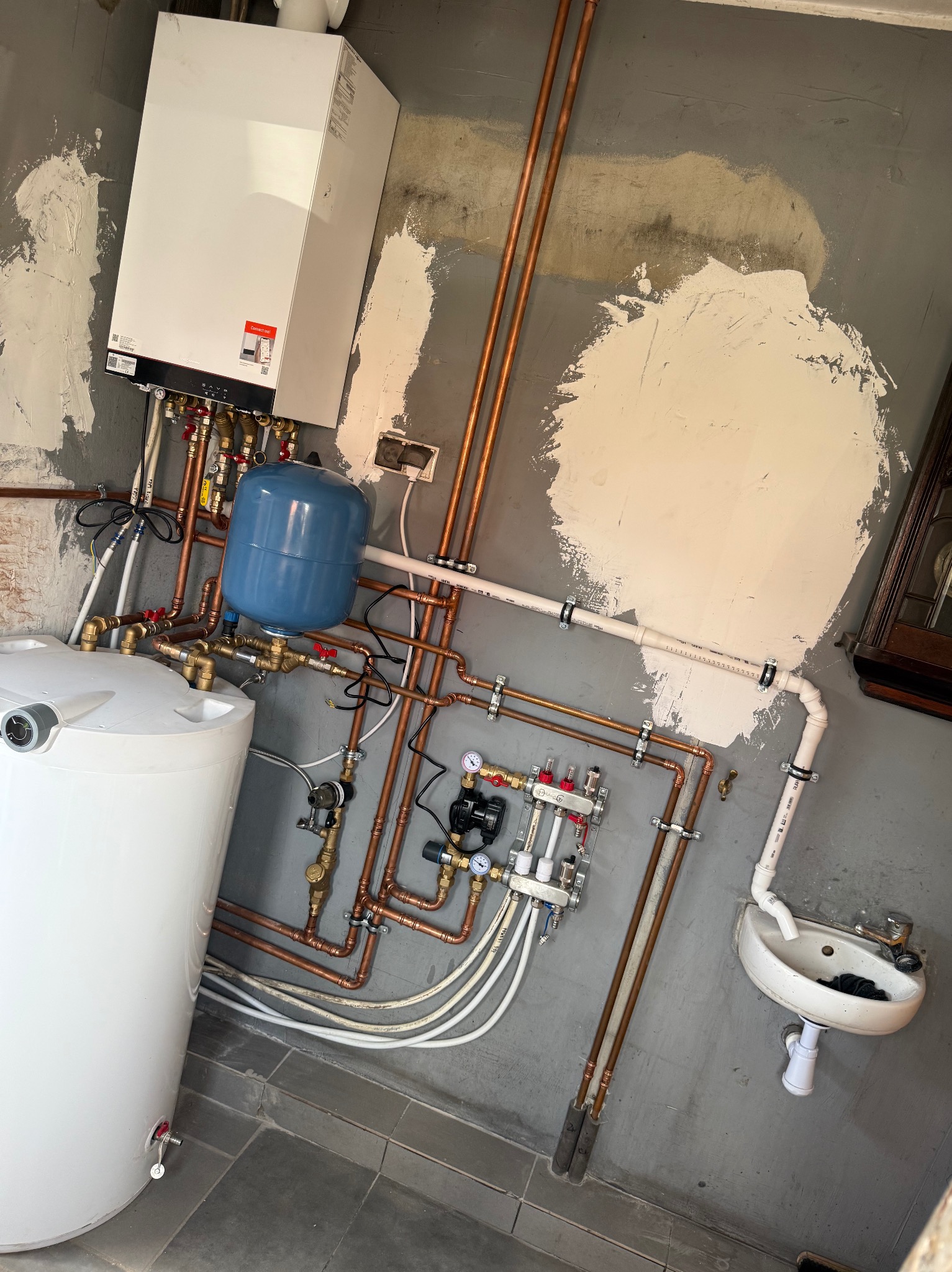 Kompleksowa instalacja grzewcza z kotłem, zasobnikiem i rozdzielaczem, rury miedziane, szare ściany w trakcie wykończenia, mała umywalka.