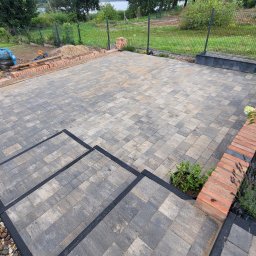 DAN-BUD Dawid Kępczyński - Ułożona kostka brukowa na tarasie z widocznymi schodami i obrzeżami, w tle teren przygotowany pod trawnik i ogrodzenie, z boku hortensja i doniczka z rośliną.