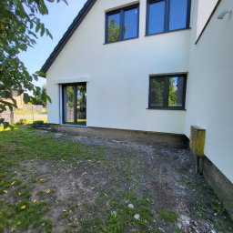 DAN-BUD Dawid Kępczyński - Świeżo otynkowany, biały dom jednorodzinny z czarną stolarką okienną i drzwiami tarasowymi, widok z boku na trawnik z resztkami gruzu.