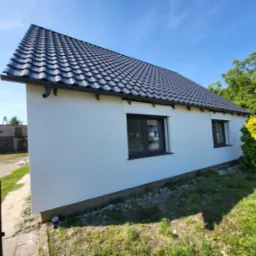 Świeżo otynkowany, biały budynek z ciemną dachówką i czarnymi oknami, widoczny z niskiej perspektywy, z fragmentem ogrodzenia po lewej stronie i zielenią w tle.