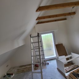 DAN-BUD Dawid Kępczyński - Poddasze w trakcie malowania: białe ściany, drewniane belki sufitowe, drabina aluminiowa, okno balkonowe, przygotowane narzędzia i materiały malarskie na podłodze.