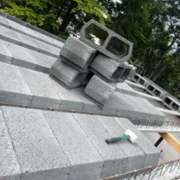 Wznoszenie ściany z szarych bloczków betonowych, widoczne zbrojenie i gumowy młotek, w tle zieleń drzew.