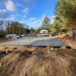 Wylana betonowa płyta fundamentowa pod budowę domu na działce z wykopami, widoczne szalunki i zbrojenia, w tle las i przyczepa kempingowa.