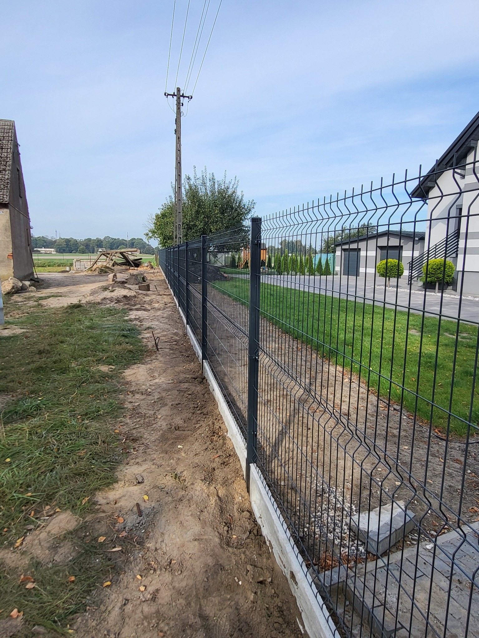 Długa, metalowa siatka ogrodzeniowa w kolorze grafitowym, zamontowana na betonowej podmurówce, oddzielająca posesję z nowoczesnym domem od terenu z budynkiem gospodarczym i słupem energetycznym.