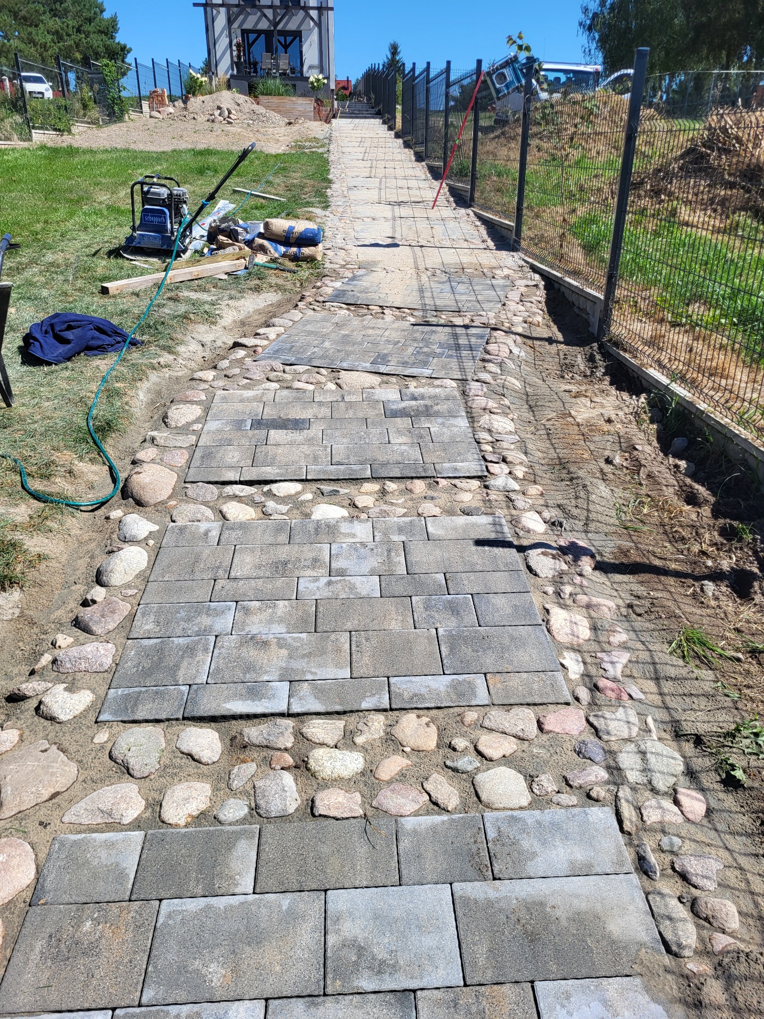 Układanie chodnika z szarych kostek brukowych, obrzeża z kamieni, w tle ogrodzenie z siatki i dom w budowie, widoczna zagęszczarka i materiały budowlane.