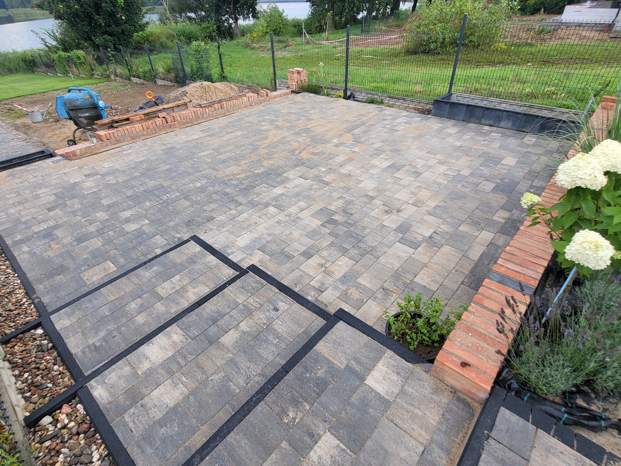 Ułożona kostka brukowa na tarasie z widocznymi schodami i obrzeżami, w tle teren przygotowany pod trawnik i ogrodzenie, z boku hortensja i doniczka z rośliną.