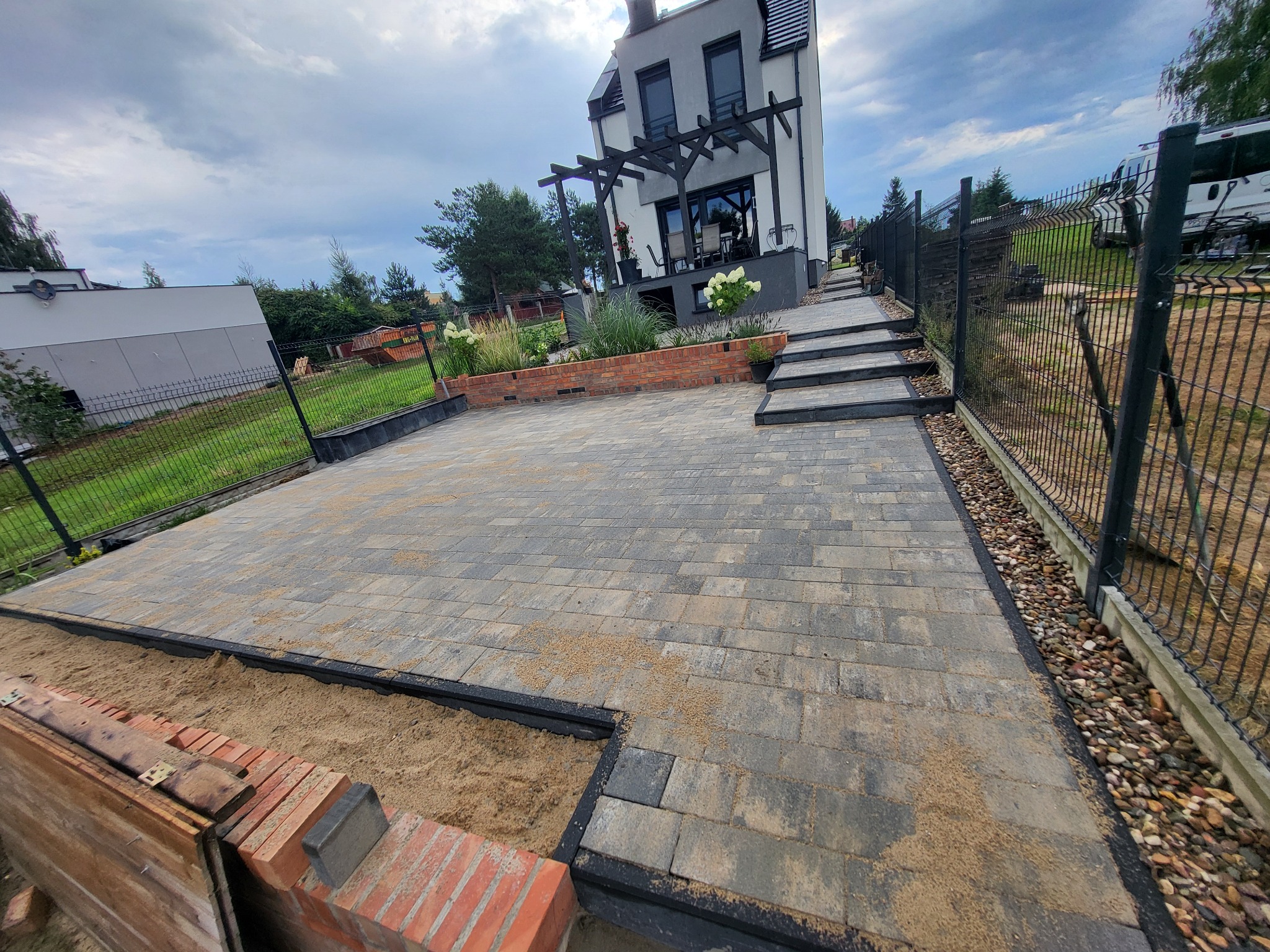 Ułożona kostka brukowa na podjeździe przed nowoczesnym domem z pergolą, widoczne obrzeża i przygotowane miejsce pod rabaty z cegły